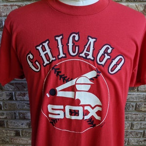 May include: Red t-shirt featuring the Chicago White Sox logo. The design includes the word "CHICAGO" in white with navy blue trim above a baseball-themed graphic and the word "SOX" in white.