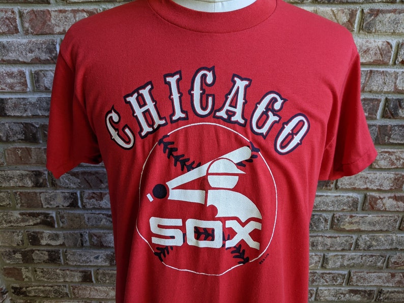 May include: Red t-shirt featuring the Chicago White Sox logo. The design includes the word "CHICAGO" in white with navy blue trim above a baseball-themed graphic and the word "SOX" in white.