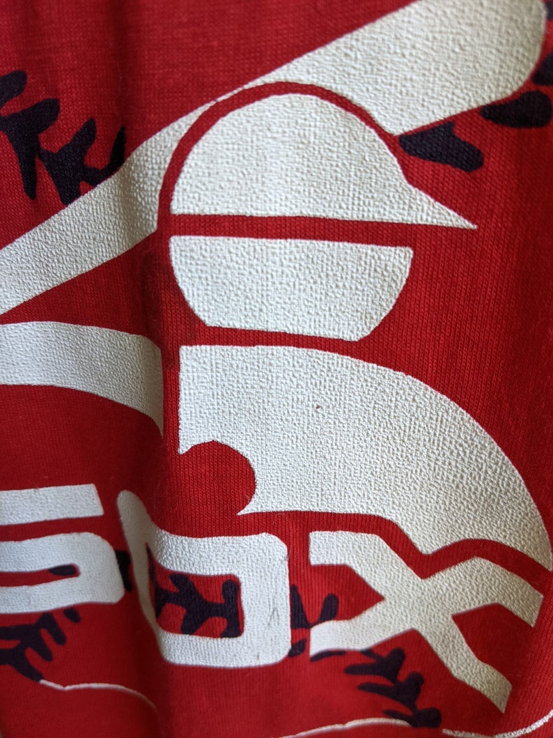 May include: Close-up of a red fabric with a large white "SOX" logo, a baseball, and baseball stitching. The logo has a textured appearance. The background is a solid red color. The image is of a sports-themed item.