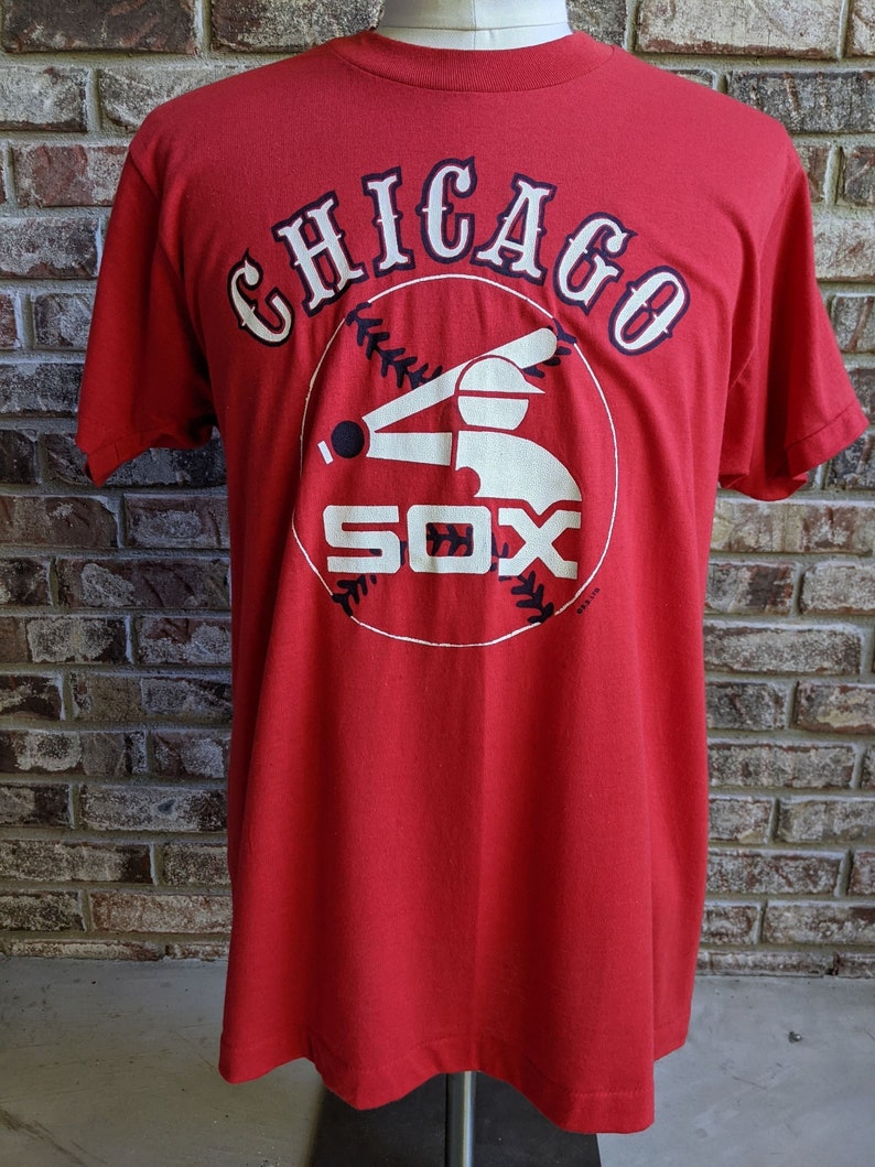 May include: Red t-shirt featuring the Chicago White Sox logo. The logo includes the word "Chicago" in white with navy blue accents, and the word "SOX" in white. A baseball and batter silhouette are also included in the design.