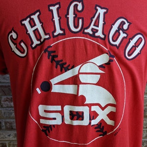 May include: Red t-shirt featuring the text "CHICAGO" arched across the top in white with navy blue trim. Below, a baseball player graphic and the word "SOX" in white. A vintage baseball tee.