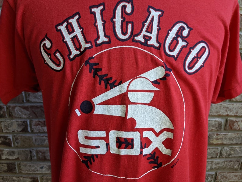 May include: Red t-shirt featuring the text "CHICAGO" arched across the top in white with navy blue trim. Below, a baseball player graphic and the word "SOX" in white. A vintage baseball tee.