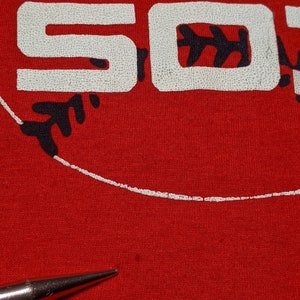May include: Close-up of a red t-shirt with the word "SO" in white, textured lettering. A black baseball design is visible. The shirt is made of a soft material.
