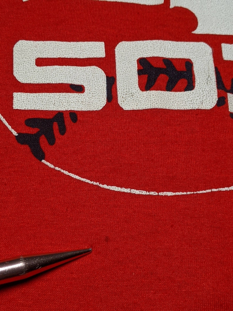 May include: Close-up of a red t-shirt with the word "SO" in white, textured lettering. A black baseball design is visible. The shirt is made of a soft material.