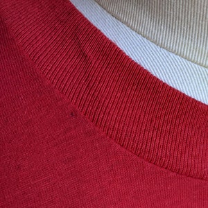 May include: Close-up of a red t-shirt collar. The collar is ribbed and a darker shade of red than the body of the shirt. The shirt is made of a soft, knit fabric. A tan and white fabric is visible in the background.