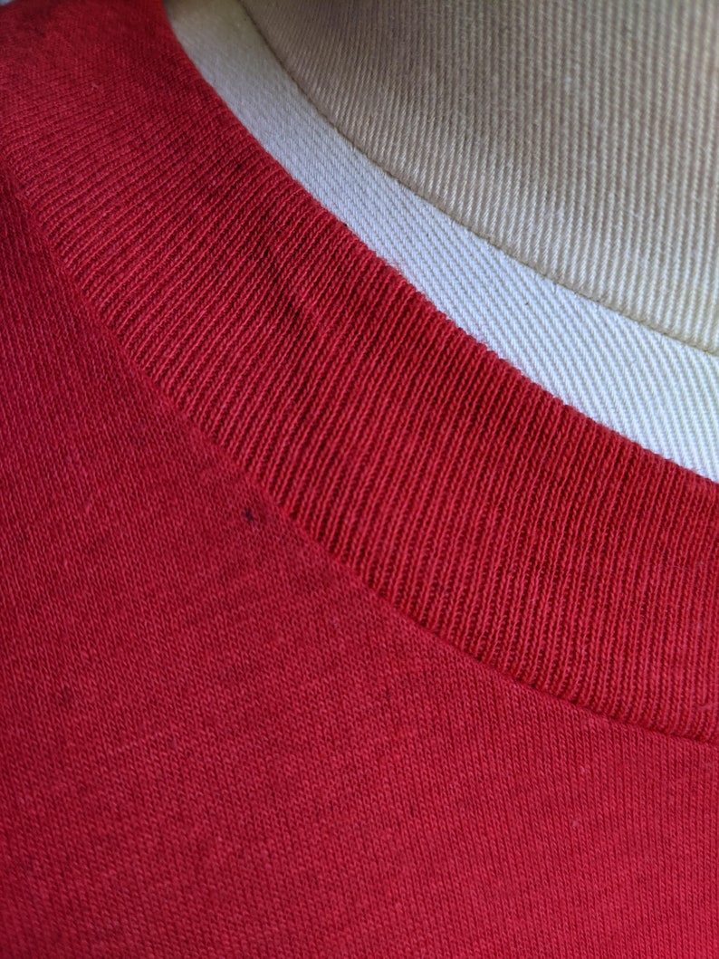 May include: Close-up of a red t-shirt collar. The collar is ribbed and a darker shade of red than the body of the shirt. The shirt is made of a soft, knit fabric. A tan and white fabric is visible in the background.