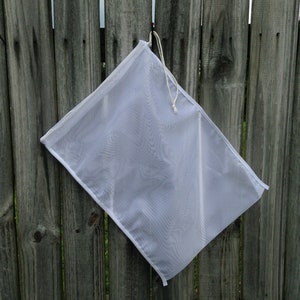 May include: A white mesh produce bag hangs on a wooden fence. The bag is empty and has a drawstring closure.
