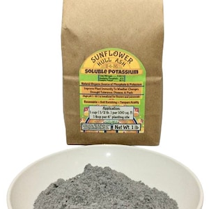 May include: A brown paper bag with a label that reads "Sunflower Hull Ash 0-6-36 Soluble Potassium". The label also includes information about the product's benefits and application instructions. A white bowl filled with gray powder is in the foreground.