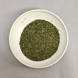 May include: A white ceramic bowl filled with dried green herbs. The herbs are finely chopped and cover the entire surface of the bowl. The bowl is centered on a gray background, providing a neutral backdrop for the food item.