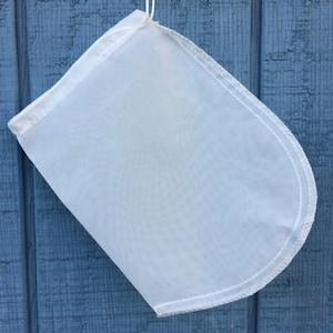 May include: A white, mesh nut milk bag with a rounded bottom and a drawstring closure. The bag is suspended by a white cord against a blue wooden background. The bag is designed for straining liquids.