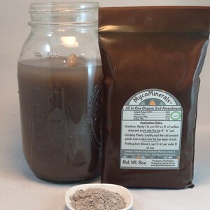 May include: A brown paper bag with the text "MycoMinerals&trade; All In One Organic Soil Amendment" and a list of ingredients and application rates. The bag is sitting on a white surface next to a glass jar filled with a dark brown liquid and a small white bowl with a gray powder.