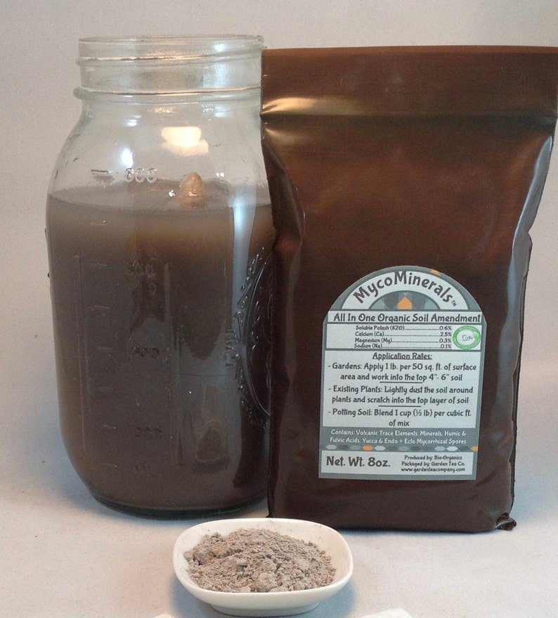 May include: A brown paper bag with the text "MycoMinerals&trade; All In One Organic Soil Amendment" and a list of ingredients and application rates. The bag is sitting on a white surface next to a glass jar filled with a dark brown liquid and a small white bowl with a gray powder.