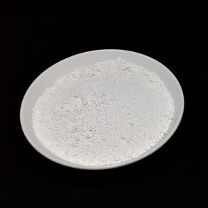 May include: A white, oval-shaped ceramic bowl filled with a fine, white powder. The bowl is centered against a stark black background, creating a high-contrast image. The powder appears granular and textured.