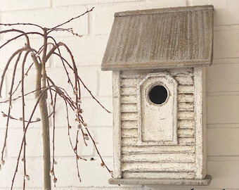 May include: A white birdhouse with a gray roof sits on a white brick wall.  The birdhouse has a small round opening for birds to enter.  There are potted plants with blue and yellow flowers in front of the birdhouse.