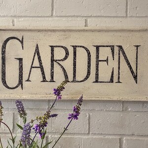 May include: White wooden sign with black lettering that reads "GARDEN". The sign is hanging on a white brick wall with purple flowers in the foreground.