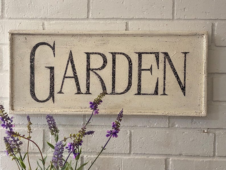May include: White wooden sign with black lettering that reads "GARDEN". The sign is hanging on a white brick wall with purple flowers in the foreground.