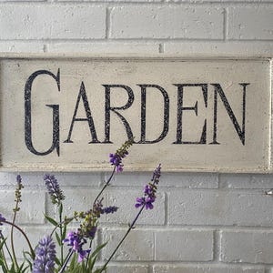 May include: A white wooden sign with black lettering that reads "GARDEN". The sign is hanging on a white brick wall. There are purple flowers in the foreground.