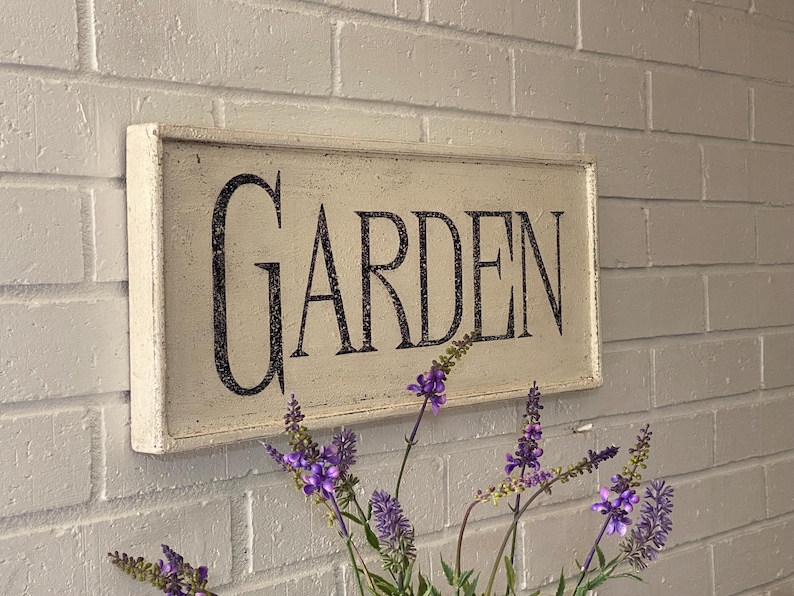 May include: White wooden sign with black lettering that reads "GARDEN". The sign is hanging on a white brick wall with purple flowers in the foreground.