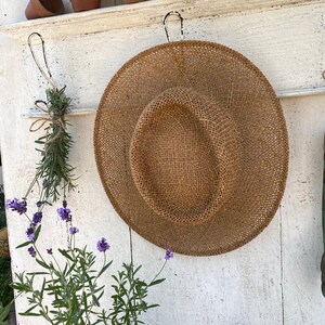 Peut inclure: Un chapeau de paille est accroch&eacute; &agrave; un mur en bois blanc avec des crochets. Le chapeau est brun et a une texture tiss&eacute;e. Il y a des plantes en pot sur une &eacute;tag&egrave;re au-dessus du chapeau.
