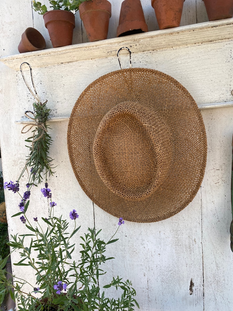 Peut inclure: Un chapeau de paille est accroch&eacute; &agrave; un mur en bois blanc avec des crochets. Le chapeau est brun et a une texture tiss&eacute;e. Il y a des plantes en pot sur une &eacute;tag&egrave;re au-dessus du chapeau.