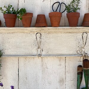 Peut inclure: Une &eacute;tag&egrave;re en bois blanc avec trois crochets, d&eacute;cor&eacute;e de plantes en pot, d'outils de jardinage et d'herbes s&eacute;ch&eacute;es. L'&eacute;tag&egrave;re est fix&eacute;e &agrave; un mur en bois blanc.
