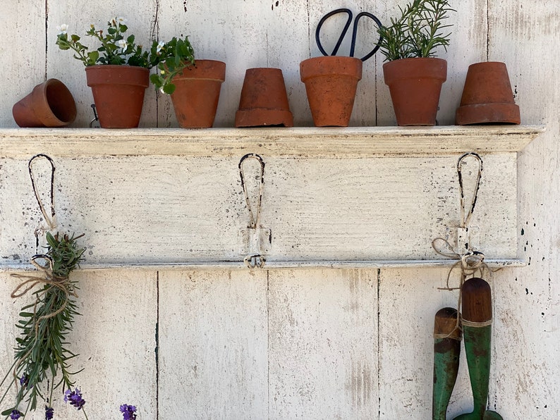 Peut inclure: Une &eacute;tag&egrave;re en bois blanc avec trois crochets, d&eacute;cor&eacute;e de plantes en pot, d'outils de jardinage et d'herbes s&eacute;ch&eacute;es. L'&eacute;tag&egrave;re est fix&eacute;e &agrave; un mur en bois blanc.