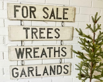 Puede incluir: Cuatro letreros rectangulares de madera blanca desgastada con letras negras cuelgan de una pared de ladrillos blancos. Los letreros dicen "FOR SALE", "TREES", "WREATHS" y "GARLANDS". Un pequeño árbol de hoja perenne está a la derecha.