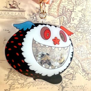 May include: A whimsical, cartoon-like keychain featuring a smiling creature with a white face, black body with red polka dots, and blue and red accents. The creature's mouth reveals a vintage map design. Attached to a gold-colored clasp.