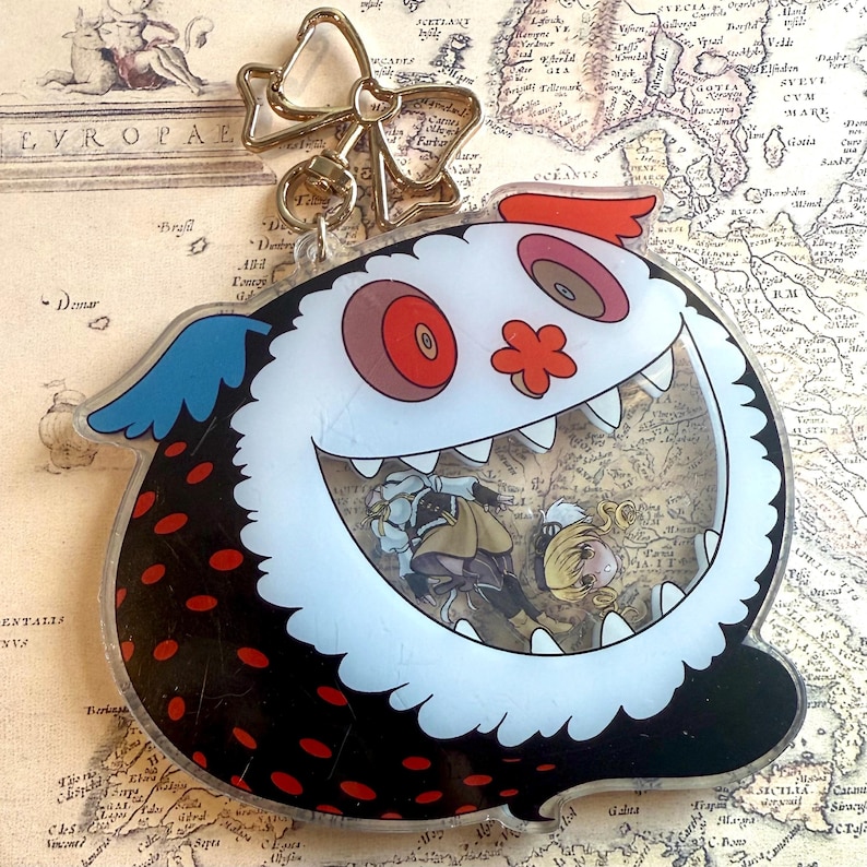 May include: A clear acrylic keychain featuring a cartoon monster design with a white face, red eyes, and a large open mouth. The monster has black spots and blue accents. Inside the monster is a picture of two anime characters. A gold-colored bow and chain are attached.