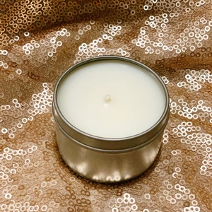 May include: A white candle in a silver tin container, sitting on a gold sequined fabric.