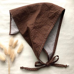 May include: A brown bonnet with a white and brown patterned design. The bonnet has a tie closure and is made of fabric. The bonnet is a traditional style, suitable for babies or children.