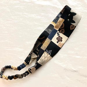May include: A black and beige headband with a Japanese-inspired pattern featuring cats, flowers, and geometric shapes. The headband is made of fabric and has an elastic band.