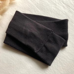 Puede incluir: Una diadema gris oscuro, doblada y colocada sobre una superficie blanca. La diadema est&aacute; hecha de un material suave y el&aacute;stico. La imagen tambi&eacute;n muestra una peque&ntilde;a cantidad de hierba decorativa beige seca.