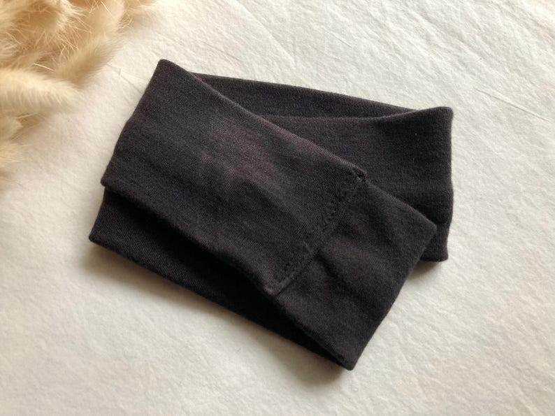 Puede incluir: Una diadema gris oscuro, doblada y colocada sobre una superficie blanca. La diadema est&aacute; hecha de un material suave y el&aacute;stico. La imagen tambi&eacute;n muestra una peque&ntilde;a cantidad de hierba decorativa beige seca.