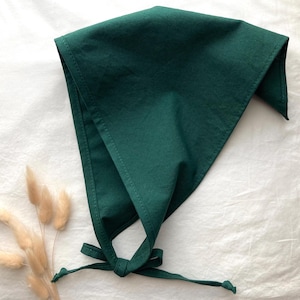Puede incluir: Un pañuelo triangular de color verde bosque oscuro sólido con bordes cosidos y dos largas cintas de tela en la base que están anudadas. La tela tiene una textura suave y mate, presentada en una forma triangular simple y plegada.