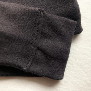 Puede incluir: Primer plano de tela gris oscuro, posiblemente una manga o dobladillo, que muestra un borde doblado y costuras detalladas. La tela parece ser un material de punto suave, con una costura visible y puntadas limpias y uniformes.