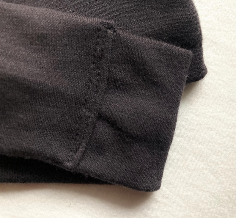 Puede incluir: Primer plano de tela gris oscuro, posiblemente una manga o dobladillo, que muestra un borde doblado y costuras detalladas. La tela parece ser un material de punto suave, con una costura visible y puntadas limpias y uniformes.