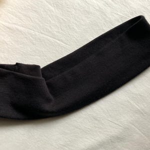 Puede incluir: Una diadema negra de tela suave, doblada para una secci&oacute;n frontal m&aacute;s gruesa. El fondo es una superficie clara y texturizada. Este accesorio es adecuado para diversas actividades. La diadema probablemente est&aacute; hecha de un material suave.