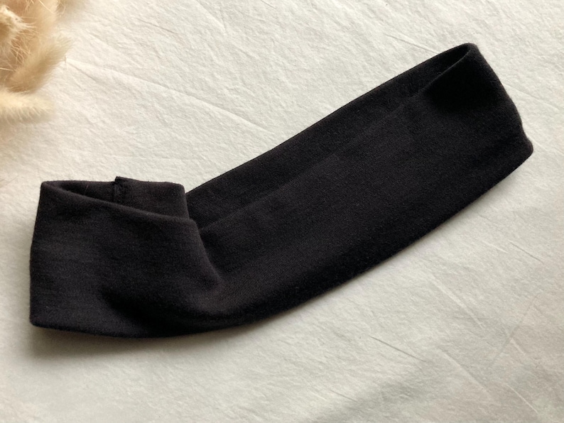 Puede incluir: Una diadema negra de tela suave, doblada para una secci&oacute;n frontal m&aacute;s gruesa. El fondo es una superficie clara y texturizada. Este accesorio es adecuado para diversas actividades. La diadema probablemente est&aacute; hecha de un material suave.