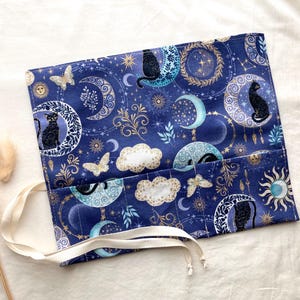Puede incluir: Un estuche de tela rectangular enrollable con fondo azul oscuro y un patrón celestial de gatos negros, lunas crecientes, soles, estrellas, nubes y mariposas. El estuche incluye una cinta de tela de color crema para cerrarlo.