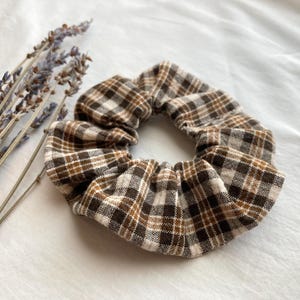 May include: A brown, white, and tan plaid scrunchie. The fabric hair accessory is circular. Dried lavender sprigs are in the background. A simple, everyday hair accessory.