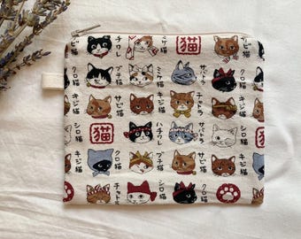 May include: A cream-colored zippered pouch featuring a repeating pattern of illustrated cat faces in various colors and styles, with Japanese text. The pouch has a small fabric loop on the side.
