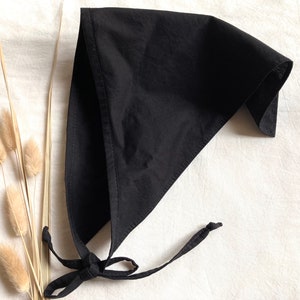 Puede incluir: Un pañuelo de algodón negro con lazos largos. El pañuelo está doblado en forma de triángulo y los lazos están atados juntos en un nudo.