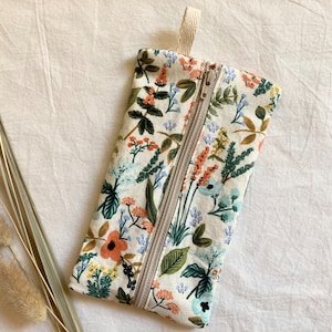 May include: A white zippered pouch with a floral print featuring various shades of blue, green, orange, and red. The pouch has a cream-colored zipper and a cream-colored fabric loop at the top.