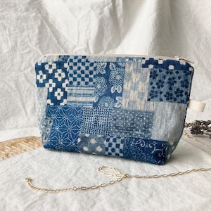 May include: A small, rectangular pouch with a patchwork design in shades of blue and white. The fabric features various geometric and floral patterns. It has a zip closure and a small loop on the side. A gold-coloured chain is attached.