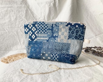 Op de afbeelding: Een kleine, rechthoekige etui met een patchwork-ontwerp in blauw- en wittinten. De stof heeft verschillende geometrische en bloemmotieven. Het heeft een ritssluiting en een kleine lus aan de zijkant. Een goudkleurige ketting is bevestigd.