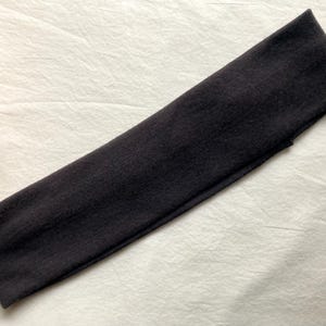 Puede incluir: Una diadema gris oscuro hecha de tela suave. La diadema es rectangular y parece el&aacute;stica. Est&aacute; sobre una superficie blanca y texturizada. La diadema es un accesorio para el cabello.