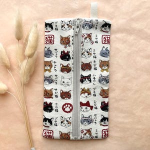 May include: A rectangular white pencil case with a zipper, featuring a repeating pattern of cat faces in various colors and Japanese text. The case has a small fabric loop at the top and a silver zipper.