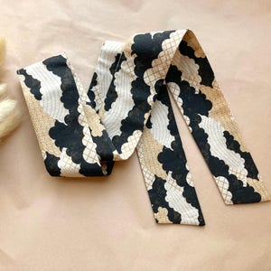 May include: A patterned fabric hair scarf in black, white, and tan. The scarf features a repeating cloud-like design with geometric accents. The scarf is laying on a light brown surface, with dried flowers in the background.