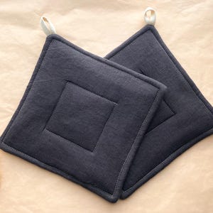 May include: Two square, dark gray pot holders with a quilted design and a loop for hanging. The pot holders are made of a textured fabric and have a contrasting white stitch around the edges.